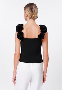 Top scurt negru cu bretele late și volănașe, design strâns pe corp, gât pătrat, purtat cu pantaloni albi largi.