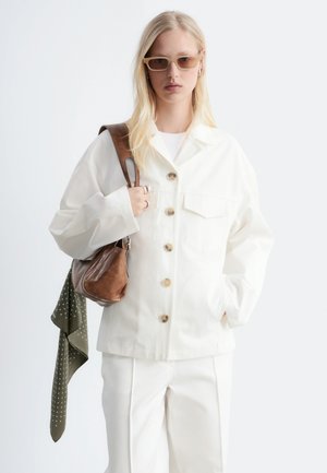 Femme blonde portant des lunettes de soleil beiges, une veste boutonnée écrue, un pantalon assorti, portant un sac à bandoulière en cuir marron avec une écharpe verte.