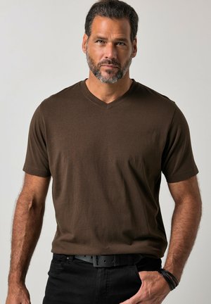 Uomo con capelli corti e scuri e barba, che indossa una maglietta marrone con scollo a V, pantaloni neri e braccialetti al polso destro, in piedi davanti a uno sfondo semplice.
