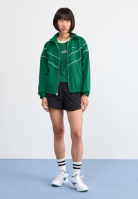 Sieviete stāv uz zilas grīdas, ģērbusies zaļā Nike jaka, zaļā kreklā, melnās šortos, baltās zeķēs ar melnām svītrām un baltās Nike sporta apavos.
