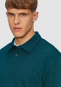 Polo shirt teal avec un col, comportant une patte de bouton et un tissu texturé. La vue en gros plan met en valeur l'ajustement et la profondeur de couleur de la chemise.
