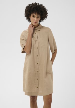 Woman with curly hair wearing beige button-up dress with collar, standing with one hand touching neck and the other in pocket.