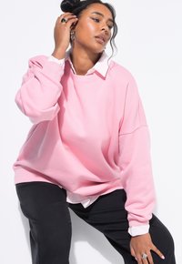 Donna con capelli scuri indossa una felpa rosa oversize sopra una camicia bianca con colletto e pantaloni neri, si tocca i capelli mentre guarda altrove.