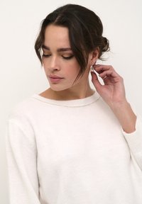 Maglione bianco con una morbida trama, caratterizzato da un sottile dettagli in argento all'collo. Il soggetto ha i capelli scuri acconciati in modo morbido. Gli orecchini d'argento vengono aggiustati.