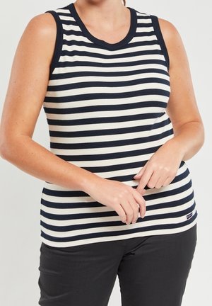 Donna che indossa una canotta a righe orizzontali bianche e nere e pantaloni neri, con le mani delicatamente intrecciate davanti.