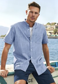 Camicia azzurro in lino a maniche corte con colletto button-down, taschino sul petto e sottili righe verticali, abbinata a pantaloni corti scuri.
