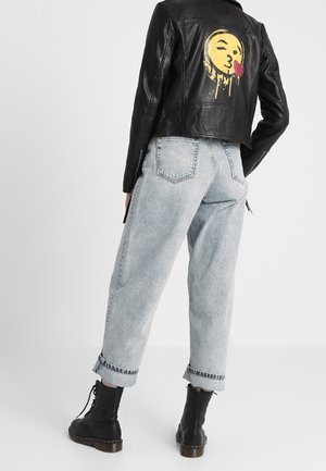 Person trägt hellblaue aufgerollte Jeans, schwarze Schnürstiefel und eine schwarze Lederjacke mit einer gelben Kussmund-Grafik auf dem Rücken.