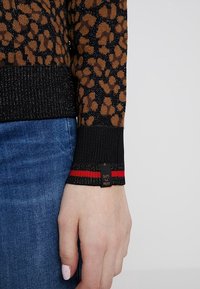 Garen gebreid sweater met een bruin en zwart luipaardprint, zwarte geribbelde boorden met rode strepen en een gestructureerde afwerking. Label zichtbaar op de boord.