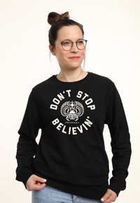 Svart sweatshirt med grafiskt tryck som har texten "DON'T STOP BELIEVIN'" och ett emblem i vitt, med ribbade mudd och rundad halsringning.