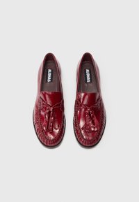 Mocassins de couro vermelho com um acabamento brilhante, apresentando um detalhe trançado e franjas decorativas na parte da frente. Sola plana, textura suave.
