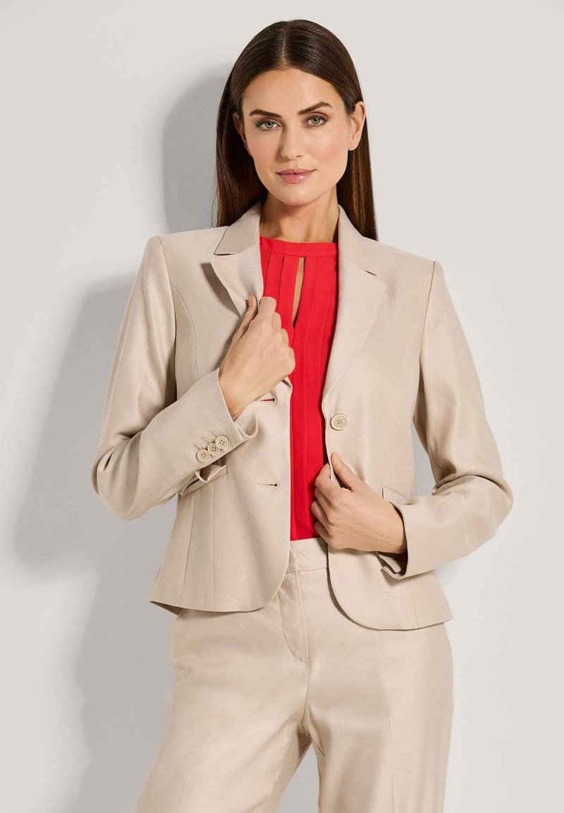 Beige op maat gemaakte blazer met knopen en gestikte details, gedragen over een felrode plooitop. Heeft een getailleerd ontwerp en een soepele textuur.