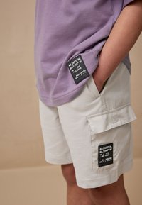 Jasnofioletowa koszulka z metką oraz kremowe szorty cargo z bocznymi kieszeniami i czarną metką na prawej kieszeni.