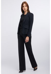 Marineblazer mit strukturiertem Design, Ein-Knopf-Verschluss, zwei Fronttaschen, kombiniert mit schwarzen, geraden Hosen und spitzen schwarzen Absätzen.