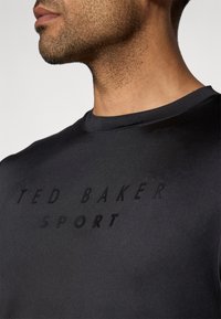 Juodas sportinis marškinėliai su apvaliu kaklu, puošti tekstūriniu spaudiniu "TED BAKER SPORT" matinės juodos spalvos ant krūtinės.