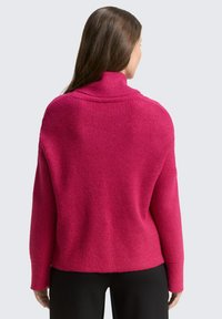 Fuchsia Pullover mit gerippter Textur, hohem Kragen und lockerer Passform. Lange Ärmel enden in weiten Bündchen. Über dunklen Hosen getragen.