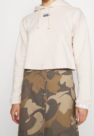 Afgekorte beige hoodie met trekkoorden en logo, gecombineerd met een bruin patroonrok met knopen aan de voorkant.