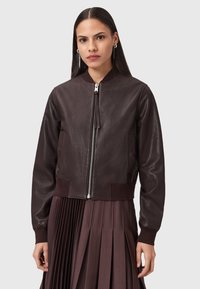 Donna che indossa una giacca bomber in pelle marrone scuro con zip argentata e una gonna plissettata coordinata, in piedi contro uno sfondo chiaro e uniforme.