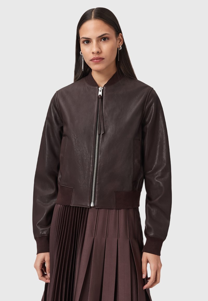 Donna che indossa una giacca bomber in pelle marrone scuro con zip argentata e una gonna plissettata coordinata, in piedi contro uno sfondo chiaro e uniforme.