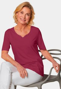 Scharlaken korte mouwen top met een V-hals en decoratieve studs bij de halslijn; gladde stof tegen witte broek, zittend op een stoel.