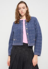 Κοντό μπλε πλεκτό ζακέτα με υφασμένη υφή, διακοσμημένο με χρυσά κουμπιά και μπροστινή τσέπη, φορεμένο πάνω από ροζ πουκάμισο και γκρι φούστα.