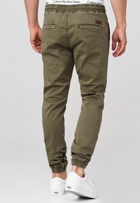 Olive green jogger pants with elastic waistband, side pockets, and elastic cuffs. Worn with white sneakers, featuring a logo on the waistband.