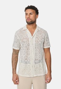 Chemise à manches courtes en blanc cassé à motif en dentelle, avec un col et une coupe ample, mettant en valeur des motifs découpés complexes.