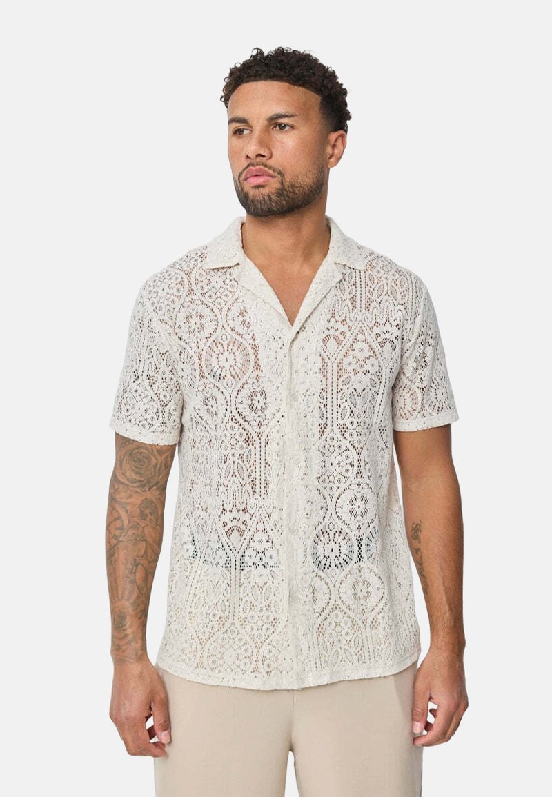 Chemise à manches courtes en blanc cassé à motif en dentelle, avec un col et une coupe ample, mettant en valeur des motifs découpés complexes.