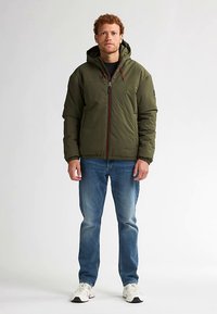 Khaki donzen jack met capuchon, ritssluiting aan de voorkant, elastische manchetten en een trekkoord. Gecombineerd met een blauwe spijkerbroek en witte sneakers, staand tegen een effen achtergrond.