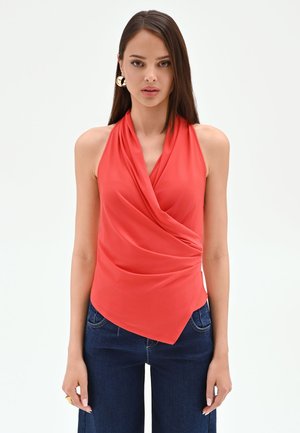 Femme aux cheveux bruns raides portant un haut rouge sans manches drapé et un jean bleu foncé, debout avec les bras détendus devant un fond blanc.