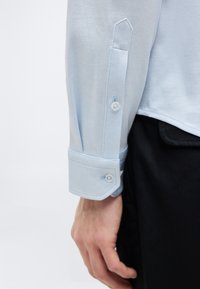 Poignet de chemise bleu clair avec patte de boutonnage portée par une personne avec main visible, associée à un pantalon noir sur fond uni.