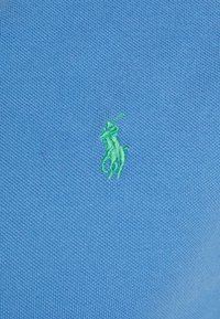 Ljusblå texturerad tyg med en liten grön broderad logotyp av en polo spelare på vänster sida. Designen är klassisk och enkel.