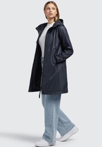 Chaqueta impermeable azul marino con capucha, cierre frontal de botones y bolsillos laterales; combinada con pantalones de mezclilla de pierna ancha azul claro y zapatillas blancas.