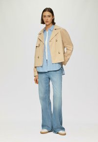 Beige cropped jas met een dubbelzijdig ontwerp, een gestreept blauw shirt eronder en lichtblauwe flared jeans, gecombineerd met witte sneakers.