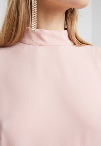 Blouse rose clair à col montant en tissu texturé ; présente un éclat élégant et subtil ainsi que des coutures visibles le long de l'encolure.