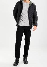 Svart puffjacka med en quiltad design, grå långärmad tröja, svarta jeans och svarta ankelstövlar. Har en huva och dragkedjedetaljer.