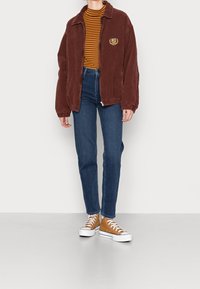 Chaqueta de pana marrón con cierre, puños acanalados, camiseta a rayas mostaza y marrón, vaqueros de tiro alto azules, y zapatillas beige con suelas blancas.