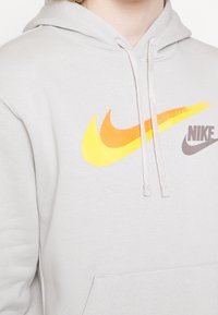 Ljusgrå hoodie med en ficka framtill, med en stor, mångfärgad Nike-logotyp i gult, orange och brunt. Dragsko i huvan.