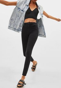 Jeans skinny de talle alto negros combinados con un top corto negro y una chaqueta sin mangas de mezclilla clara, completados con sandalias negras.