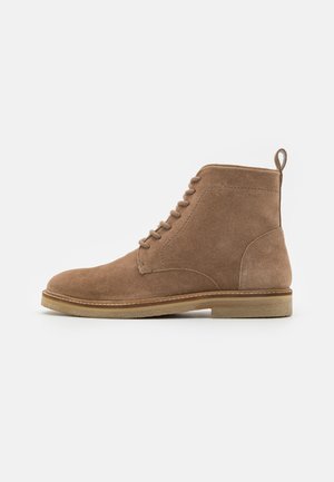 Brown suede ankle boot with round toe, lace-up design, and small pull tab at the back. Features a contrasting light-colored sole.