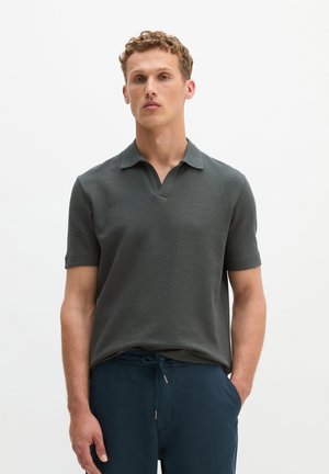 Junger Mann mit kurzen lockigen Haaren, der ein dunkelgraues Polohemd und marineblaue Hose mit Kordelzug trägt, steht vor einem schlichten weißen Hintergrund.