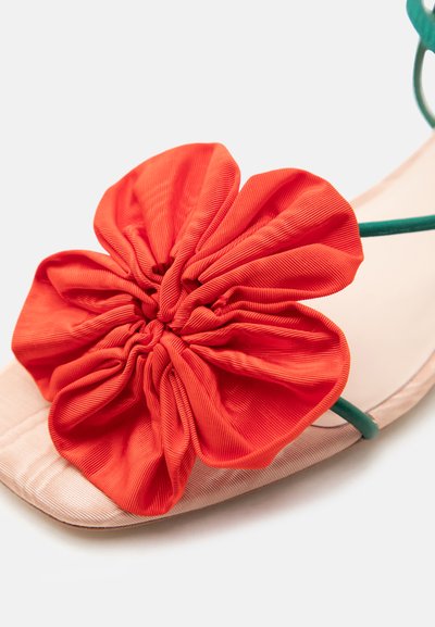 Platte sandaal met een grote rode stoffen bloem als versiering en dunne groene bandjes op een lichtbeige zool.