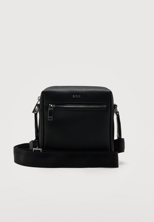 Melna teksturēta ādas plecu soma ar priekšējo rāvējslēdzēja kabatu, metāla logotipu un regulējamu auduma siksnu.