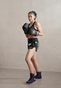Boxer wearing black gloves and a black sports bra, paired with black shorts featuring a green pattern. Footwear is blue with red accents.