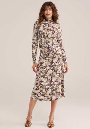 Femme portant une robe midi à manches longues et col montant avec un motif cachemire multicolore et des sandales à talons ouvertes sur un fond uni.