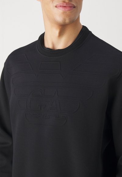 Felpa nera con design del logo in rilievo sul petto, colletto rotondo e texture liscia. Vista completa della parte superiore del torso.