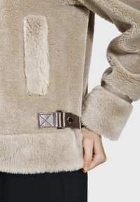 Beige wollen jas met een zachte textuur, voorzien van een zijzak en een bruine band met een zilveren metalen gesp aan de onderkant.