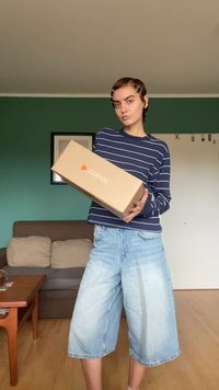 Brown cardboard box with Zalando logo; person wears a navy striped sweater and light blue wide-leg shorts, standing in a room.