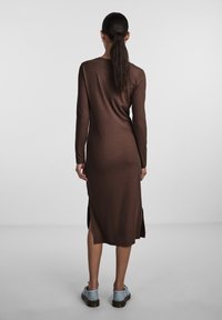 Abito in maglia a coste marrone con maniche lunghe e spacco laterale. Indossato con scarpe azzurro chiaro, presenta una silhouette aderente e una texture liscia.