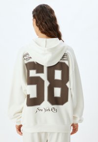 Weißer Kapuzenpullover mit einer großen braunen "58" auf der Rückseite, darüber "New York City" in dekorativen Schriftzügen und grafischen Akzenten. Angenehme Textur.