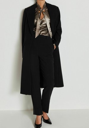 Femme portant un pantalon noir ajusté, un long manteau noir, un chemisier bronze brillant avec un nœud à l'avant et des talons pointus noirs, debout contre un mur blanc.